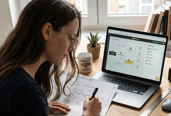 Femme de profil avec lunettes et cheveux longs travaillant sur un bureau organisé, remplissant un arbre généalogique papier à côté d'un ordinateur affichant un logiciel de généalogie, avec un classeur d'archives