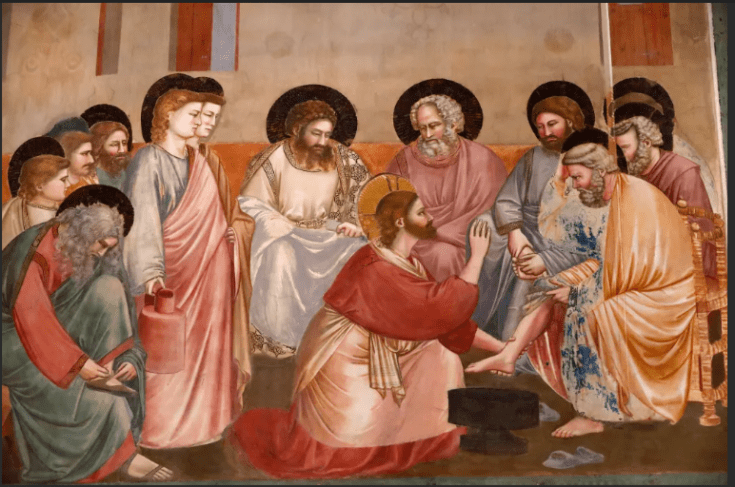 Une fresque médiévale du peintre italien Giotto di Bondone. La scène représente le Christ agenouillé, un halo doré autour de la tête, lavant le pied d'un disciple assis (probablement saint Pierre). D'autres apôtres, tous avec des halos circulaires, sont rassemblés autour, regardant avec solennité, y compris un disciple assis à gauche retirant ses sandales. L'arrière-plan montre une architecture simple dans des tons ocre et terre.