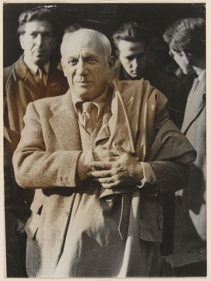 Photographie d'époque de Pablo Picasso en costume, les mains croisées sur un vêtement, entouré d'autres personnes.